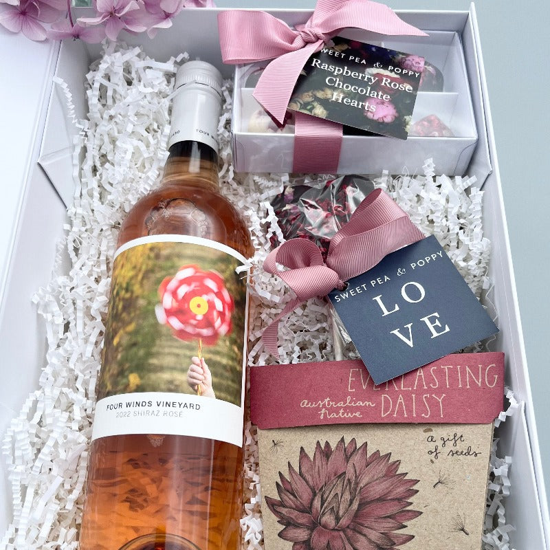 A gift hamper with rosé wine, a gift box of handmade chocolate hearts, a chocolate heart lollipop and a gift of seeds card in a white hamper box.