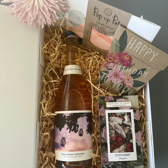 A birthday gift hamper with rosé wine, dark chocolate bar, happy birthday flower seeds and a pop up flower pot, in a white hamper box. 