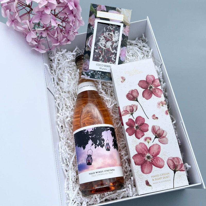 A gift hamper with sparkling rosé wine, a coco berry chocolate bar, a and a Myrtle and miss diffuser set in a white hamper box.