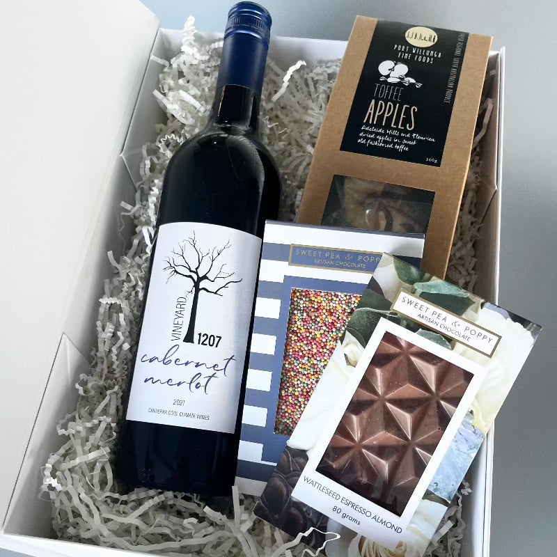 A gift hamper with red wine, a sprinkle chocolate bar, a wattleseed espresso almond chocolate bar and dehydrated toffee apples in a white hamper box.