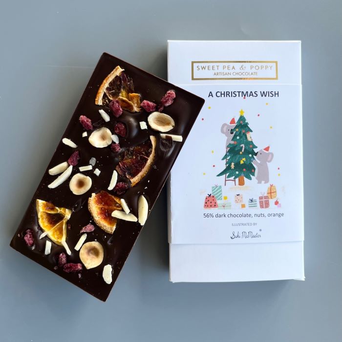 A dark chocolate handmade chocolate bar with almonds, hazenuts, cherry and orange sitting on top of a packaged and illustrated A Christmas Wish artisan chocolate bar