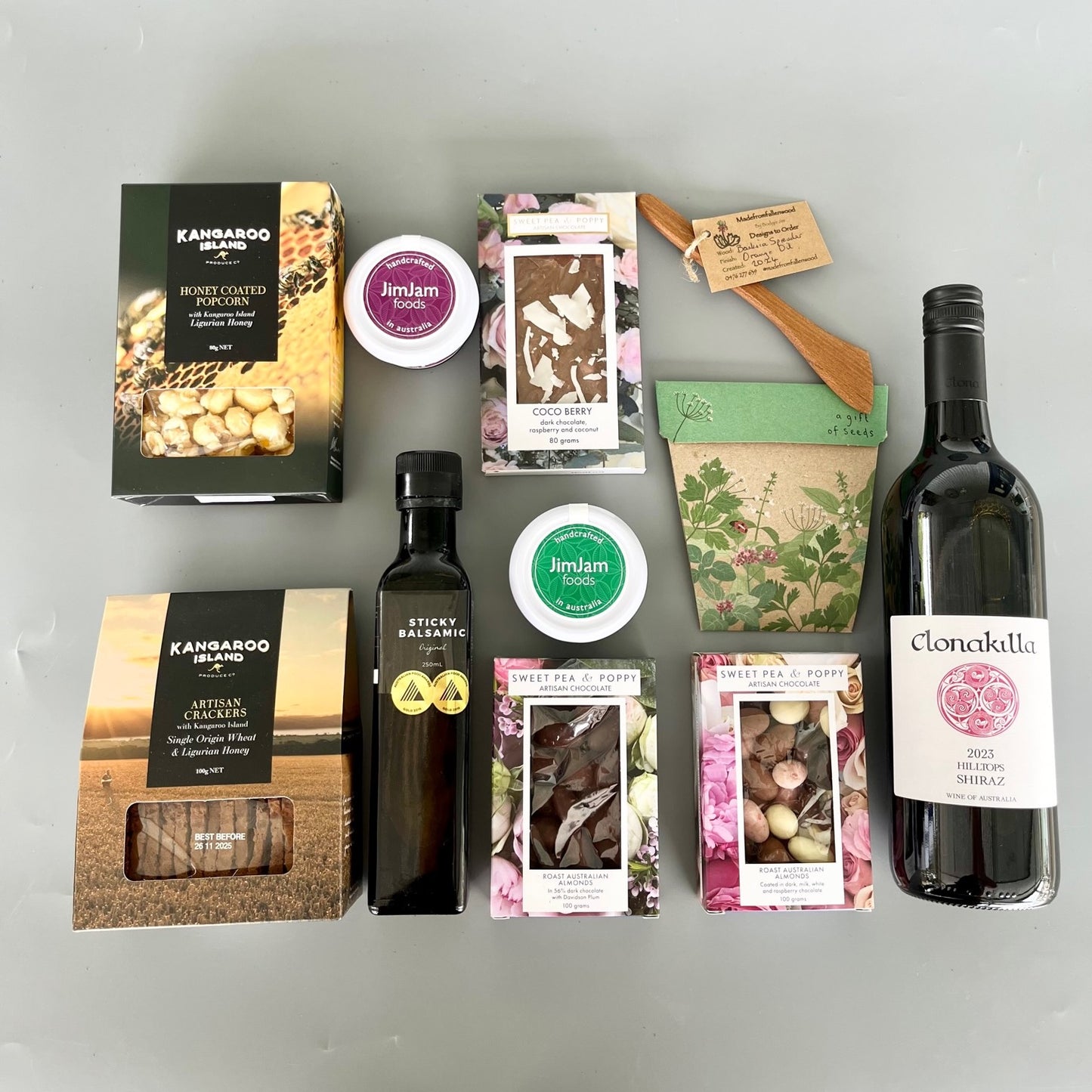 A flatlay gift hamper with wine, chocolate, wooden server, gourmet pastes, balsamic vinegar, crackers, popcorn and a gift of seeds 