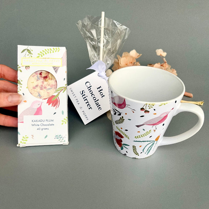 White mug with floral design, hot chocolate stirrer, and packaging on a gray background