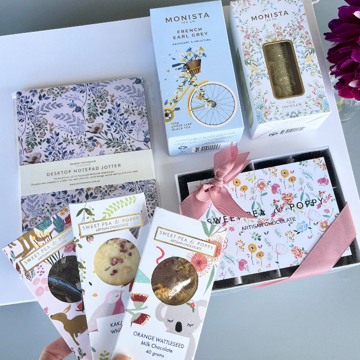 Collection of Monista tea, Sweet Pea & Poppy chocolate and Bespoke Letterpress journal on a light surface with a hand holding a box.