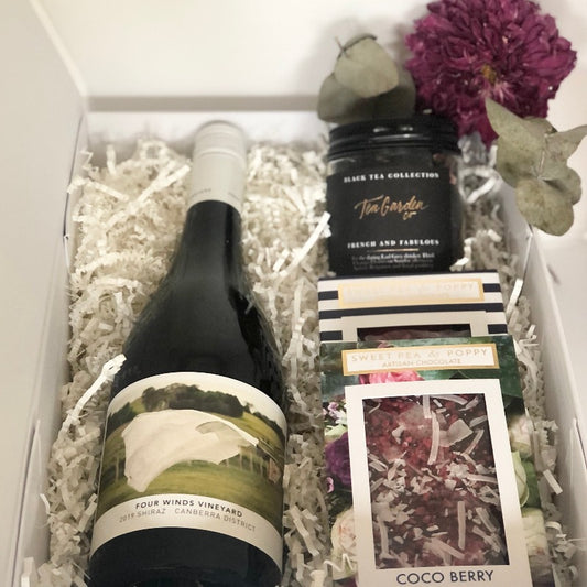 A gift hamper with red wine, a Coco Berry chocolate bar, a milk chocolate sprinkle chocolate bar and tea in a white hamper box. 