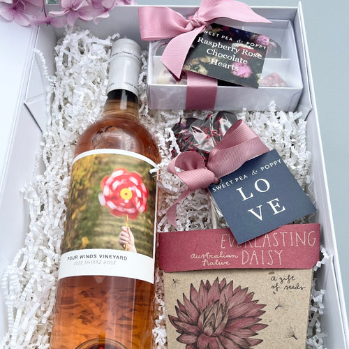 A gift hamper with rosé wine, a gift box of handmade chocolate hearts, a chocolate heart lollipop and a gift of seeds card in a white hamper box. 