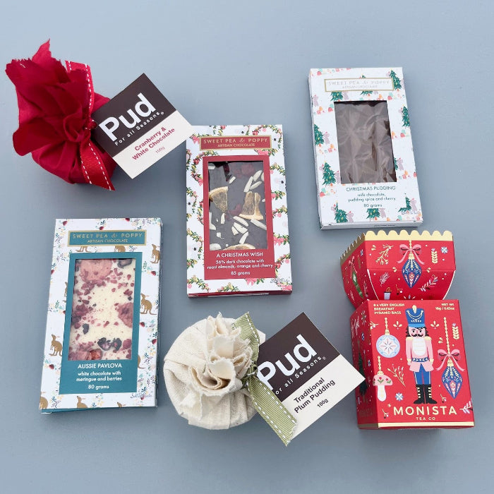 A flatlay on a grey background with three Sweet Pea & Poppy artisan chocolate bars, two individual Christmas puddings and a festive themed Christmas tea gift box