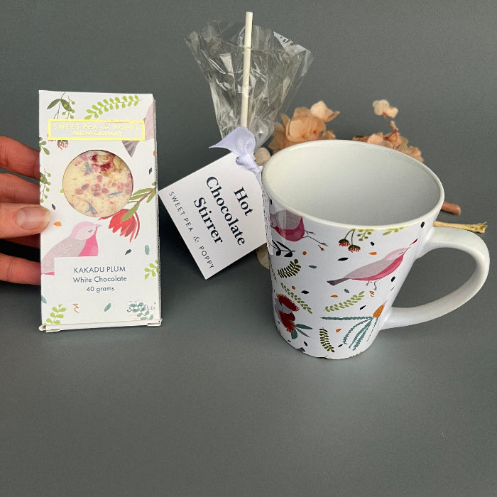 Decorative mug with floral design, hot chocolate stirrer, and packaging on a gray background