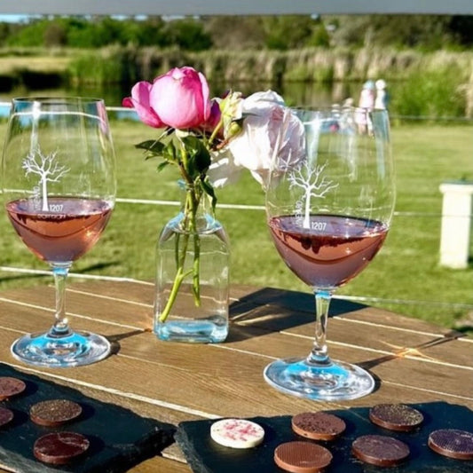 Two glasses of wine with chocolate platters and a vase of flowers on a wooden table outdoors.